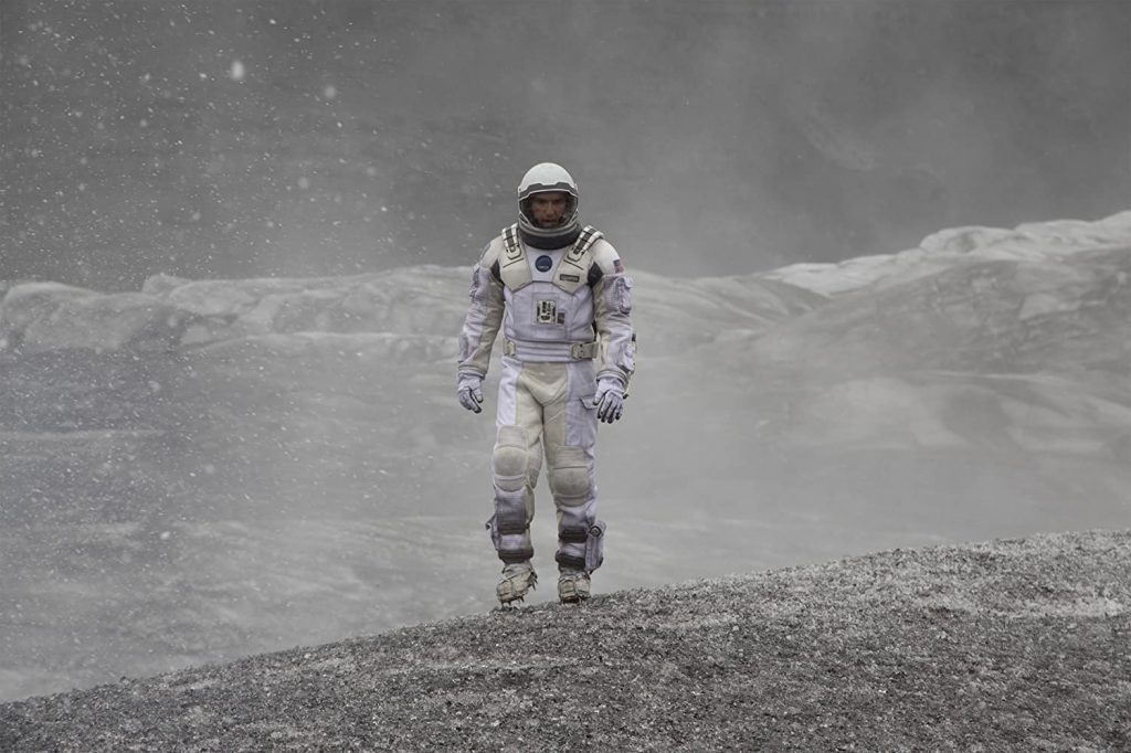 Astronauta en traje espacial caminando sobre terreno árido y rocoso de otro planeta, con montañas al fondo