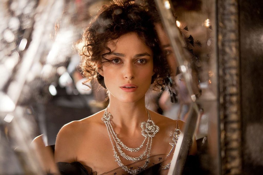 Mujer con cabello rizado recogido, collar de perlas y broche decorativo, maquillaje elegante, en ambiente luminoso