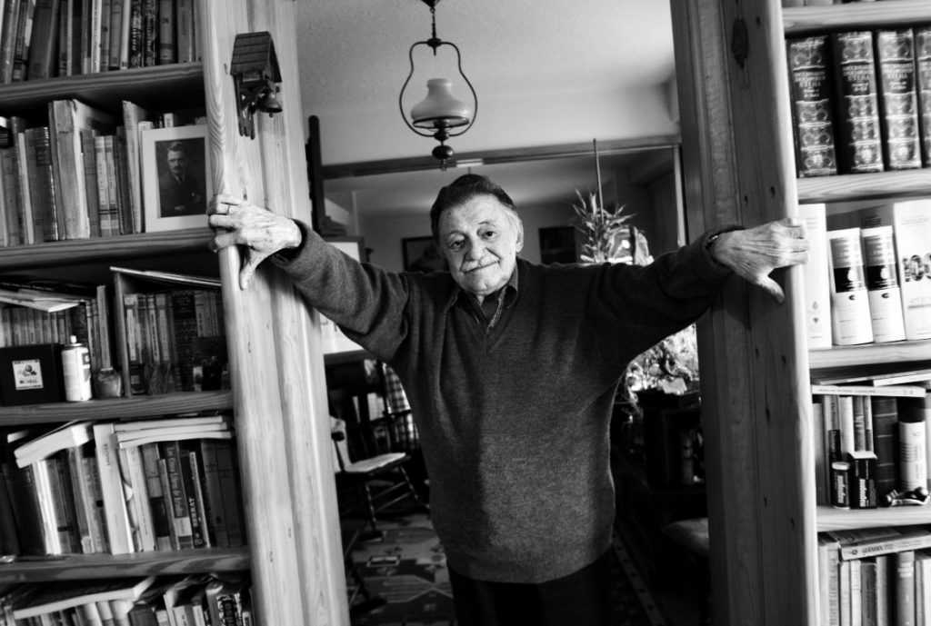 "Fake News": El poder de la mentira Mario Benedetti en su biblioteca, brazos extendidos entre estantes de libros, fotografía en blanco y negro