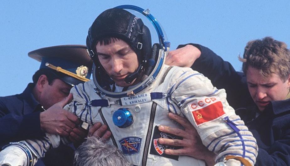 Cosmonauta Sergei Krikaliov siendo asistido tras descenso de nave espacial, con técnicos en traje oscuro a ambos lados