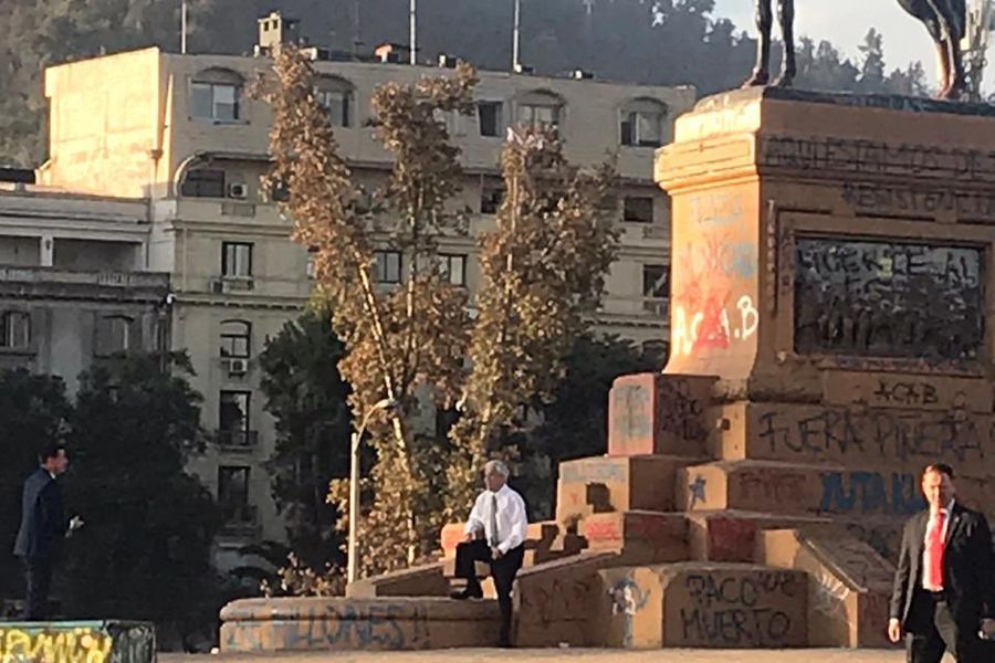 Monumento público con figura central de bronce, edificios de apartamentos al fondo y árbol dorado iluminado por luz