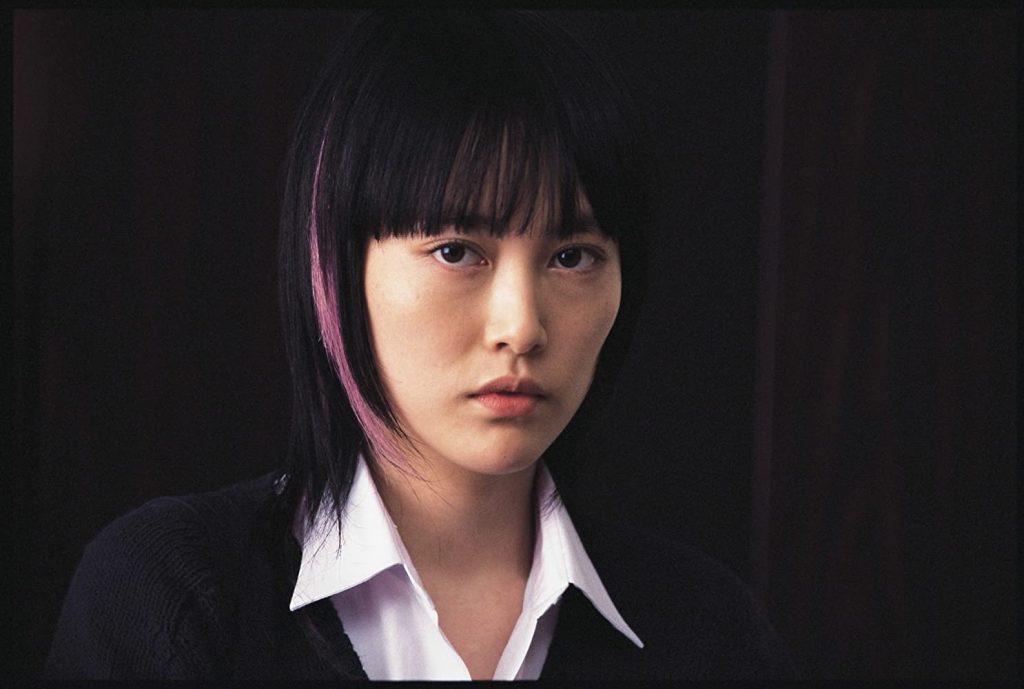 Mujer joven con cabello oscuro y flequillo, viste blazer negro con camisa blanca, expresión seria contra fondo oscuro