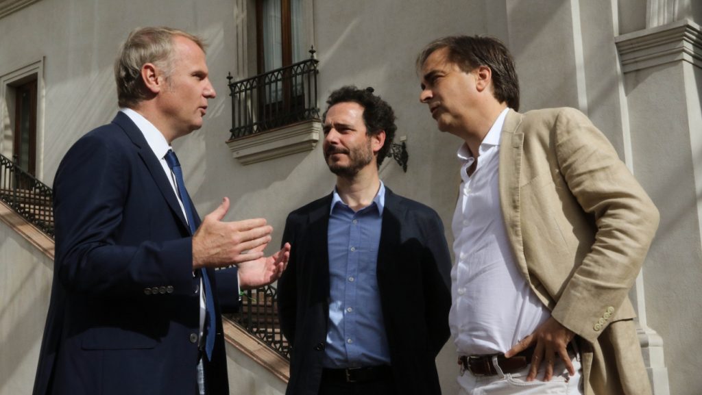 Tres hombres conversan en una terraza exterior, uno de ellos viste traje oscuro mientras los otros dos llevan camisas