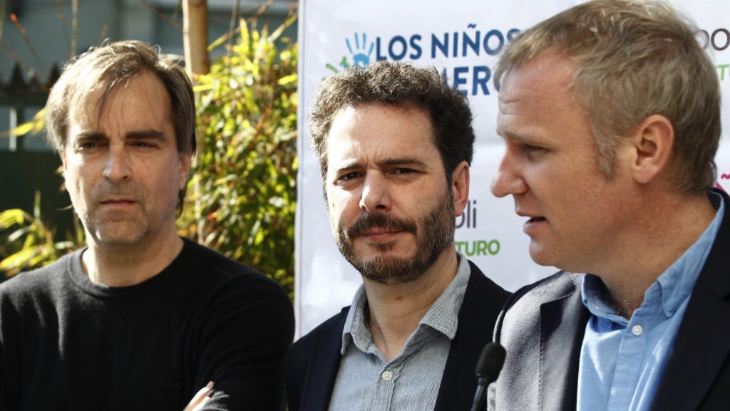 Hambruna se escribe así: La estética del "Estado Evopoliano" Tres hombres en evento público: uno de perfil a la izquierda, dos conversando al centro y derecha frente a banner
