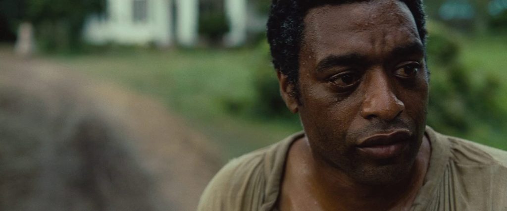 Hombre afroamericano con expresión seria en camisa clara, fondo rural desenfocado, escena de drama histórico