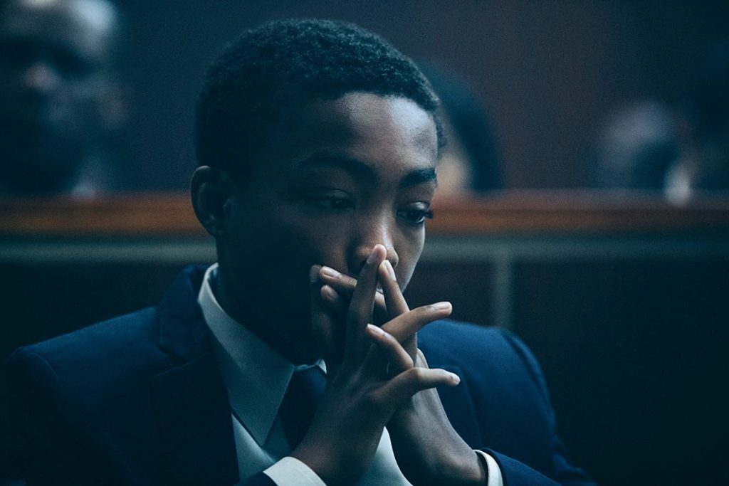 Hombre afroamericano en traje azul, sentado en sala de tribunal, con expresión pensativa y mano en el rostro