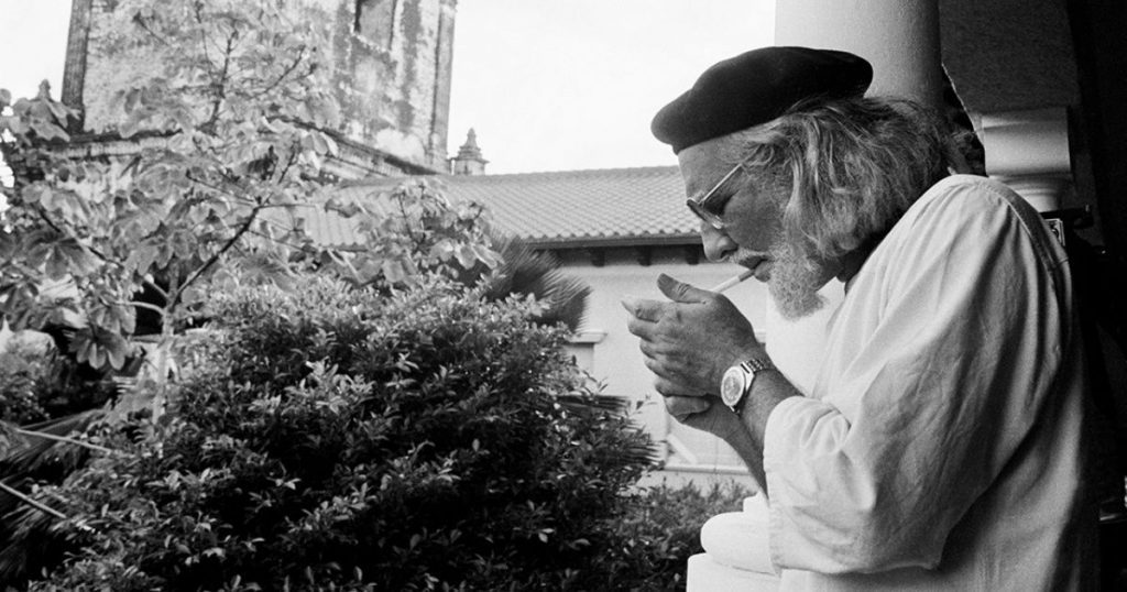 Ernesto Cardenal, poeta y sacerdote nicaragüense, observa plantas en un jardín con edificios de fondo