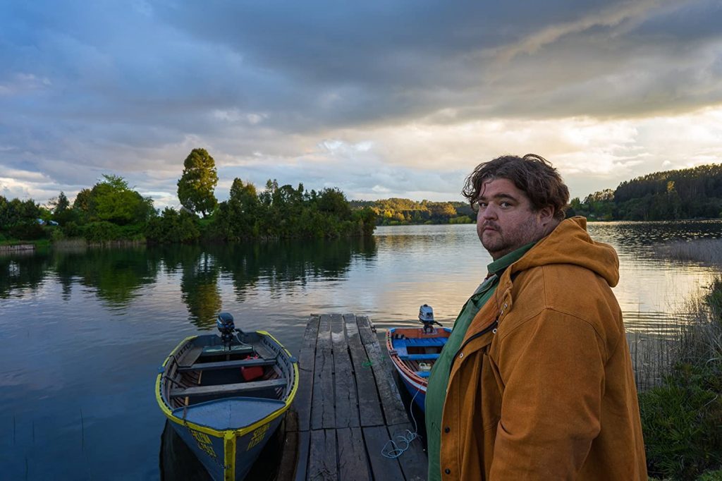 Hombre en chaqueta marrón junto a botes de madera en muelle al atardecer, frente a lago tranquilo