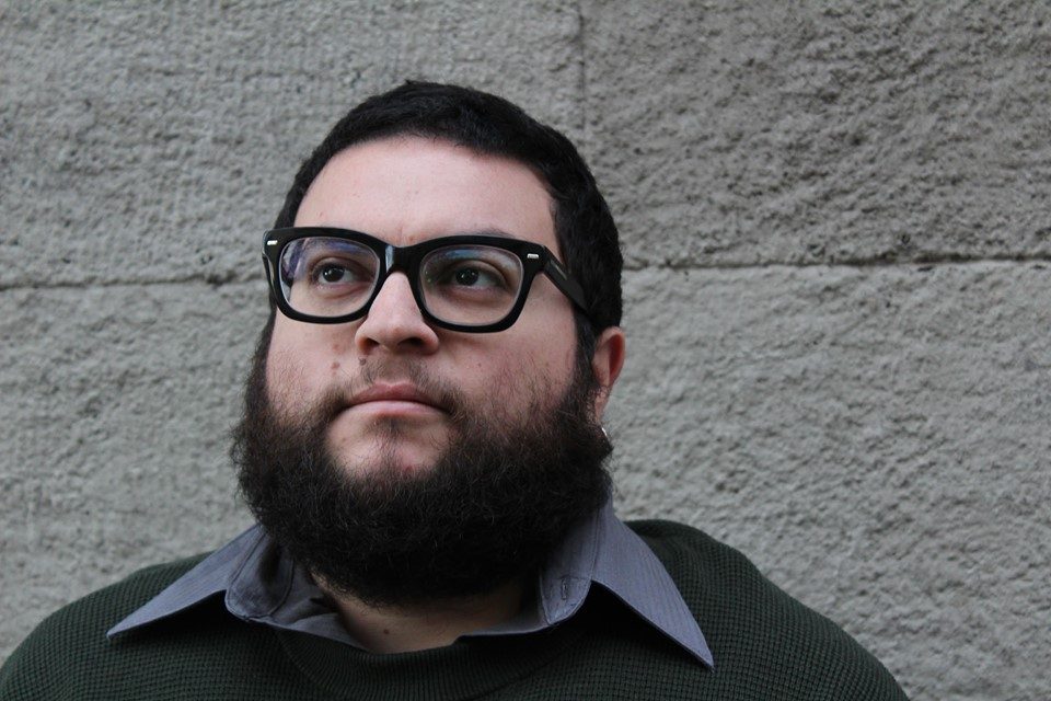 Simón Soto, hombre de barba y gafas, frente a pared gris, retrato en primer plano