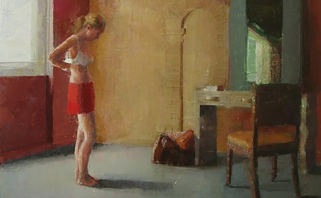 Mujer desnuda de pie en habitación con paredes ocres y rojas, junto a silla de madera y espejo, en escena íntima