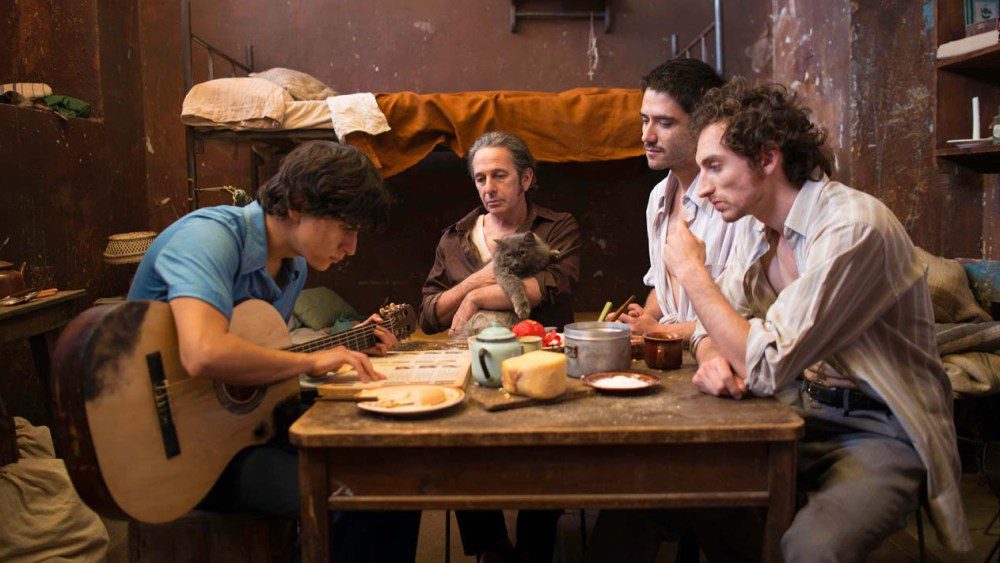 Cuatro personas alrededor de una mesa de madera comparten comida y conversación en un espacio interior cálido y austero
