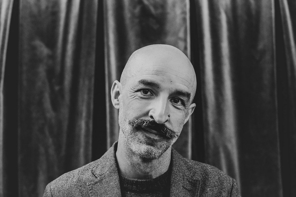 "Intemperie", de Jesús Carrasco: Los paisajes de lo inhumano Retrato en blanco y negro de Jesús Carrasco, escritor español, de frente con bigote y fondo de cortinas