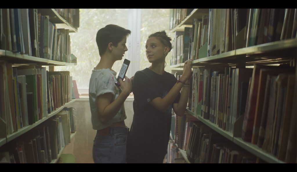 A propósito de "La jauría": ¿Qué imaginarios representan a la comunidad LGBT+ en la ficción audiovisual? Dos jóvenes conversan entre estantes de libros en una biblioteca, ambiente íntimo de literatura y diálogo