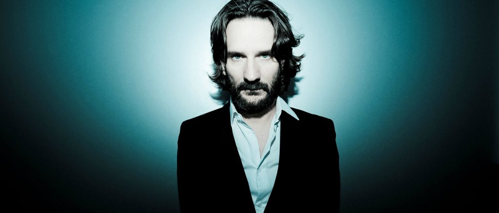 Frédéric Beigbeder, escritor francés con barba y cabello oscuro, retrato sobre fondo azul turquesa