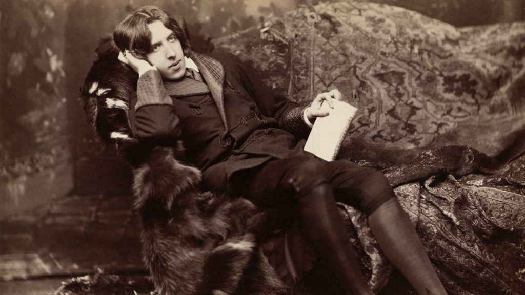Oscar Wilde recostado leyendo un documento, vistiendo traje oscuro, en fotografía sepia vintage