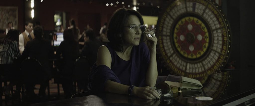 Mujer de mediana edad en interior oscuro con reloj de pared al fondo, expresión pensativa, escena de drama