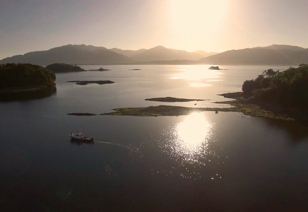 Bahía al atardecer con embarcaciones, islas boscosas y montañas al fondo bajo luz dorada reflejada en el agua