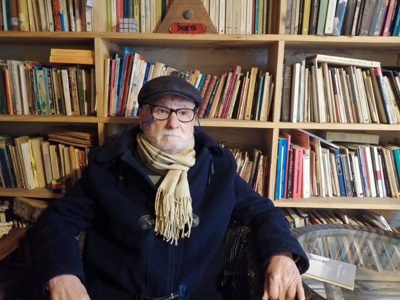 Hernán Miranda Casanova, escritor chileno, sentado frente a estantería con libros, usando gafas y bufanda