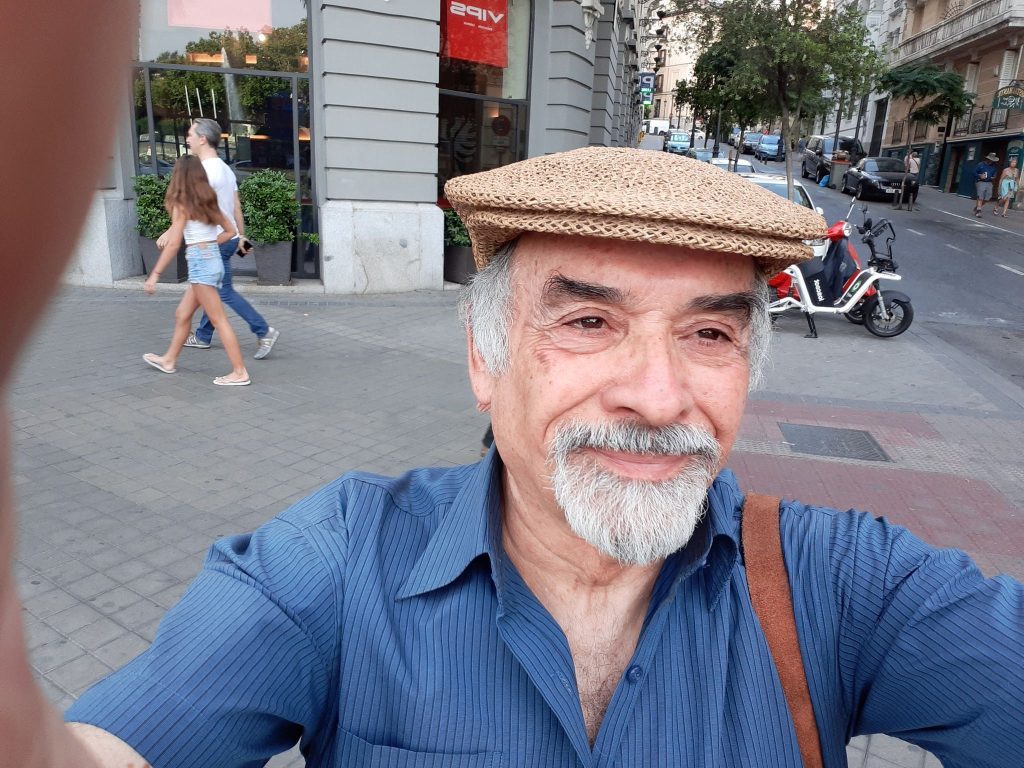 Escritor Juan Mihovilovich sonríe a la cámara en una calle urbana, luciendo gorra de lino y camisa azul