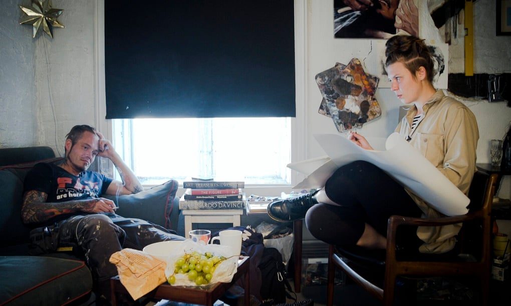 Dos personas en una sala conversando junto a una guitarra y frutero. Luz natural de ventana. Ambiente íntimo y artístico