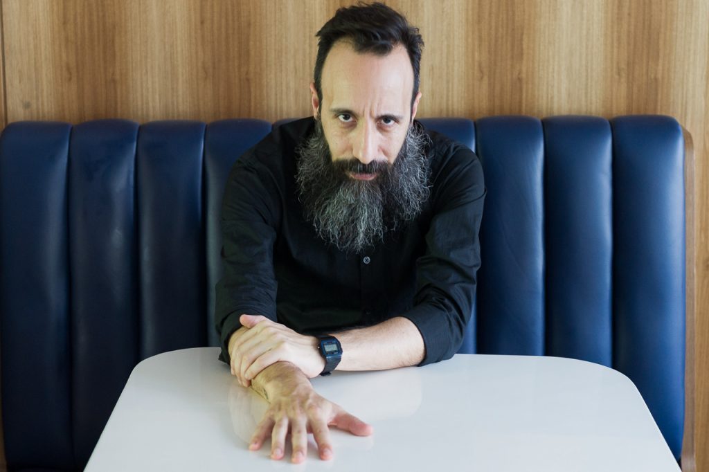 Marcelo Vera, escritor con barba oscura, apoya su mano sobre una mesa blanca frente a un sofá azul