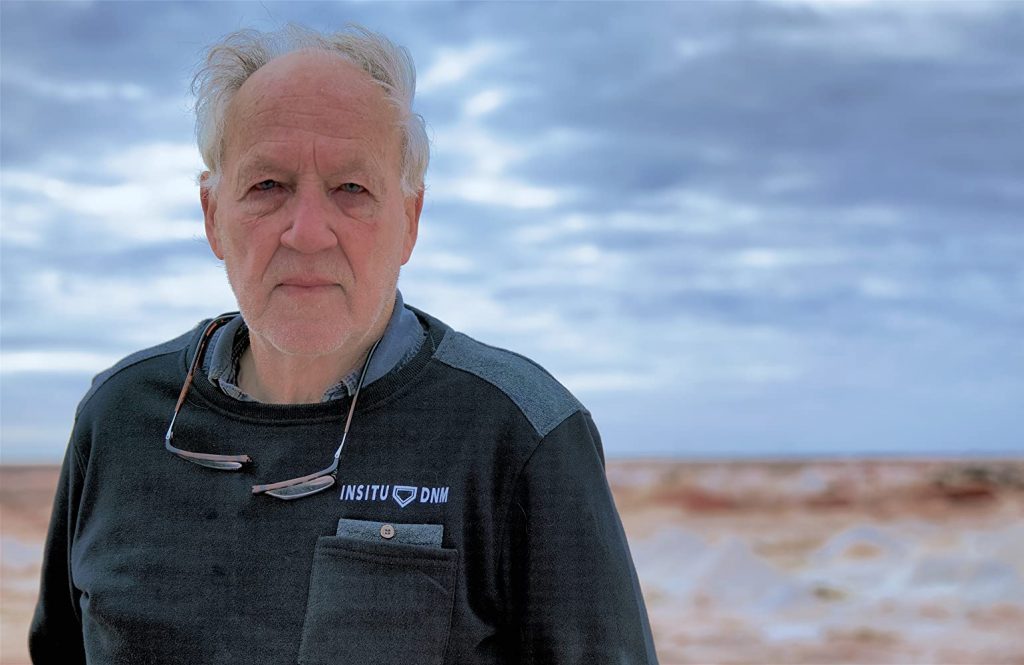 Werner Herzog en paisaje desértico, retrato de perfil frontal con cielo nublado de fondo