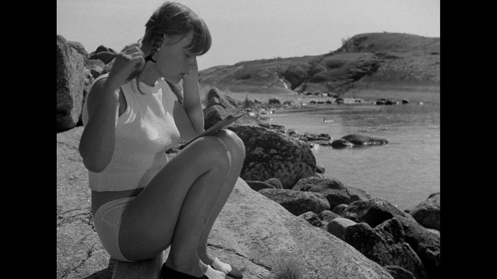 Mujer joven sentada en la playa sosteniendo binoculares, paisaje rocoso al fondo, fotografía en blanco y negro