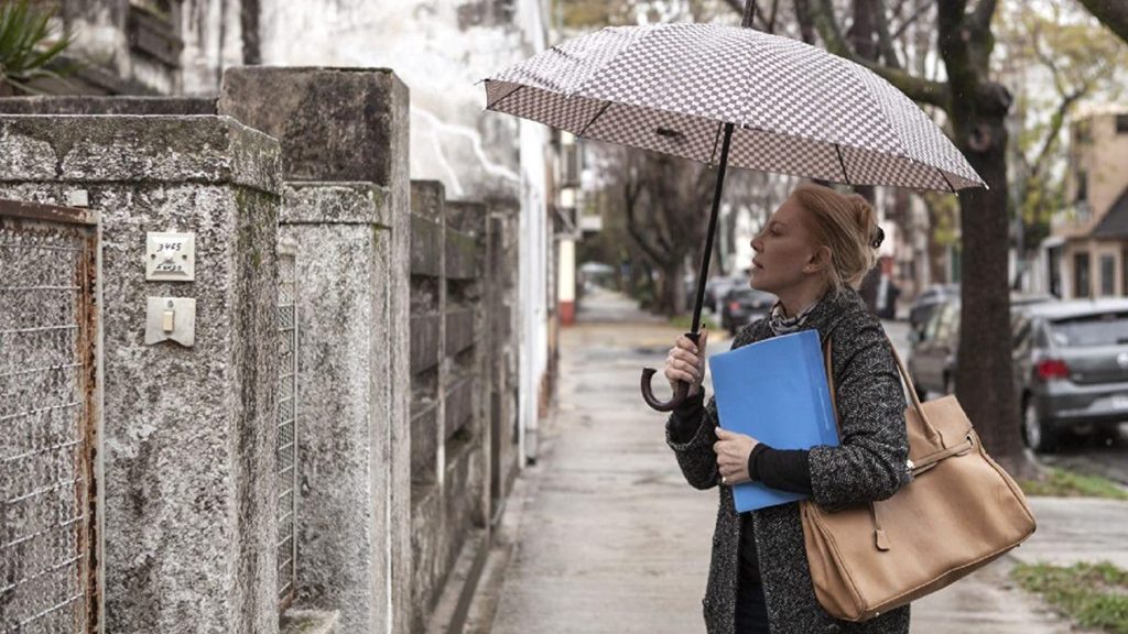 "Crímenes de familia": El thriller de Netflix donde brilla Cecilia Roth Mujer sosteniendo paraguas y carpeta bajo lluvia en calle empedrada, junto a pared de piedra blanca