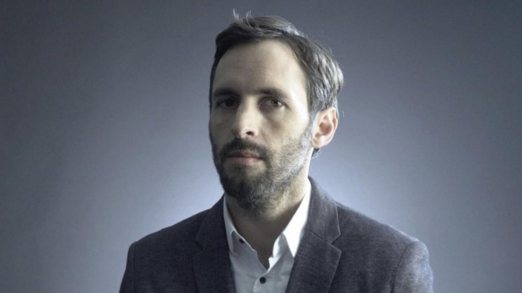 Declararse un idiota: Una necesidad vital para entender la esencia de Chile Daniel Matamala, hombre de mediana edad con barba, camisa oscura, fondo gris neutro, expresión seria y contemplativa