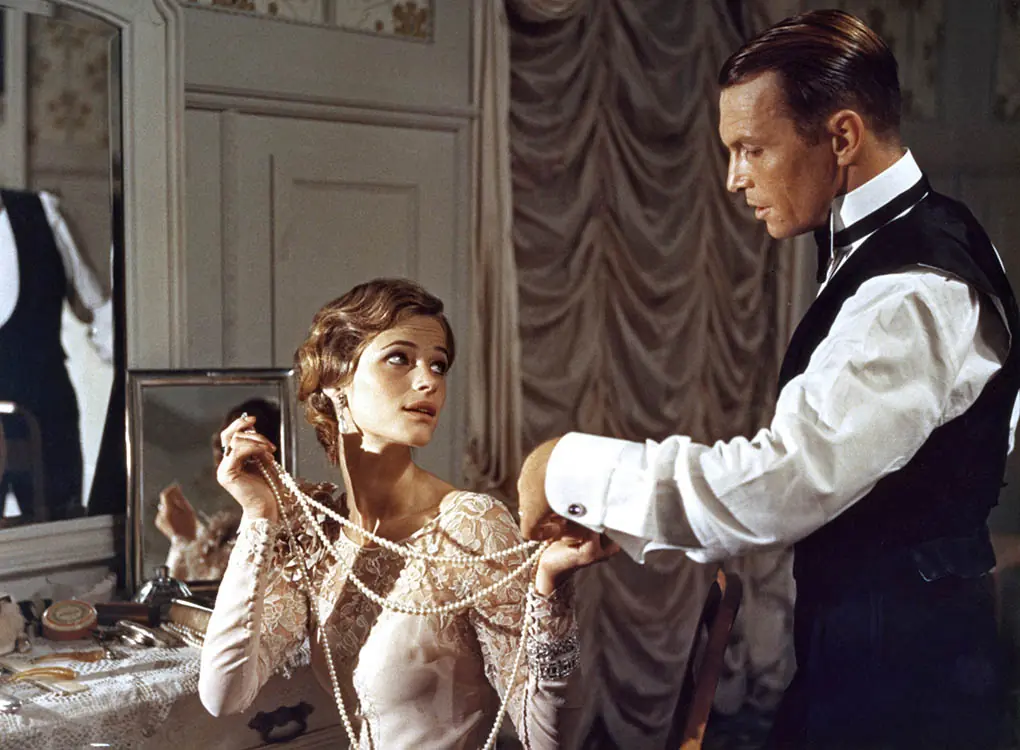 Escena de época: hombre de traje sostiene los brazos de una mujer joven en interior aristocrático con cortinas elegantes