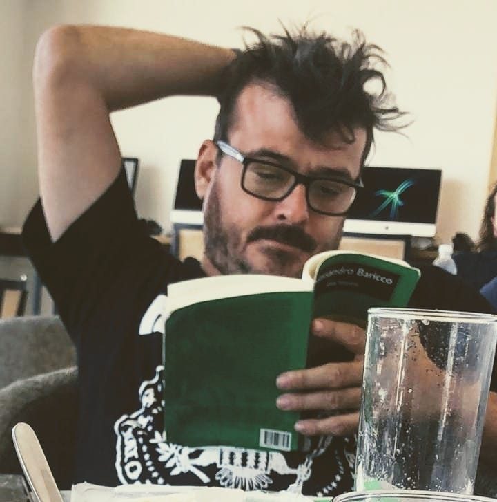 Juan José Podestá leyendo un libro de portada verde, con gafas y gesto contemplativo en su escritorio