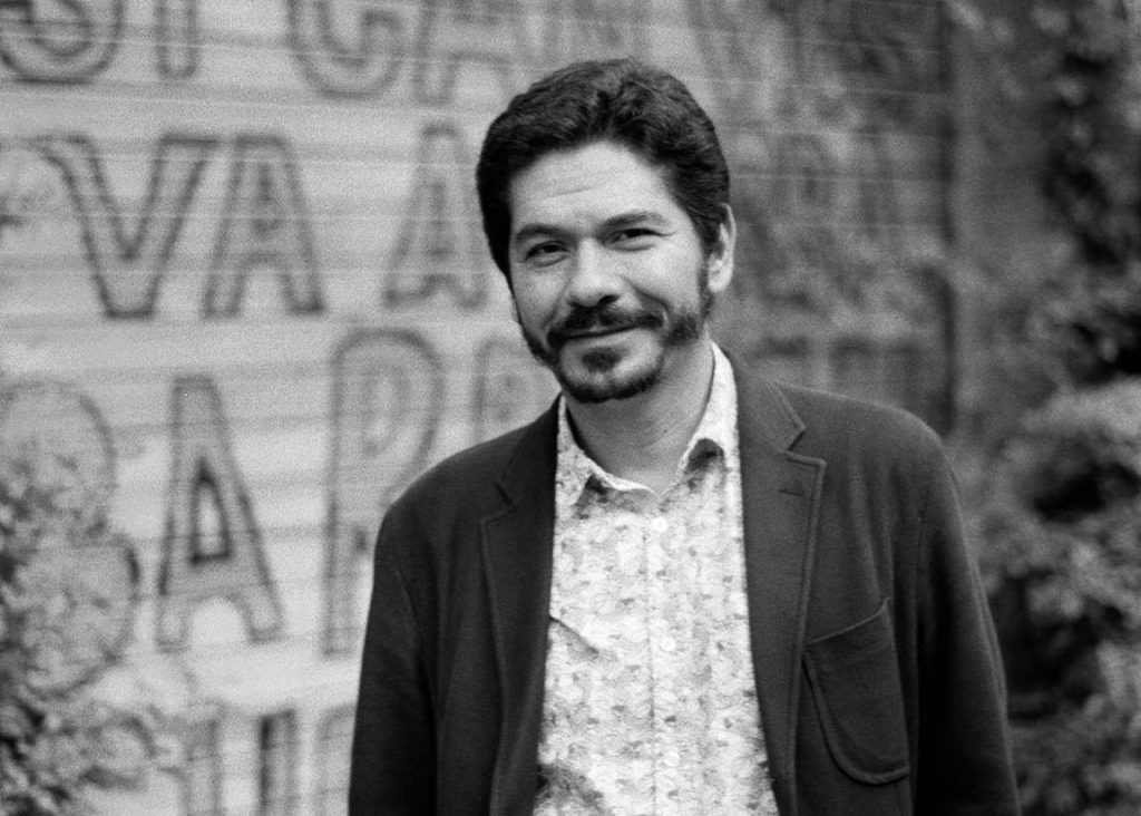 Retrato en blanco y negro de Juan Pablo Villalobos, escritor mexicano, frente a un muro con texto parcialmente visible
