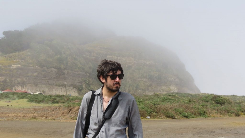 Hombre con gafas de sol y chaqueta gris frente a montaña brumosa y vegetación verde