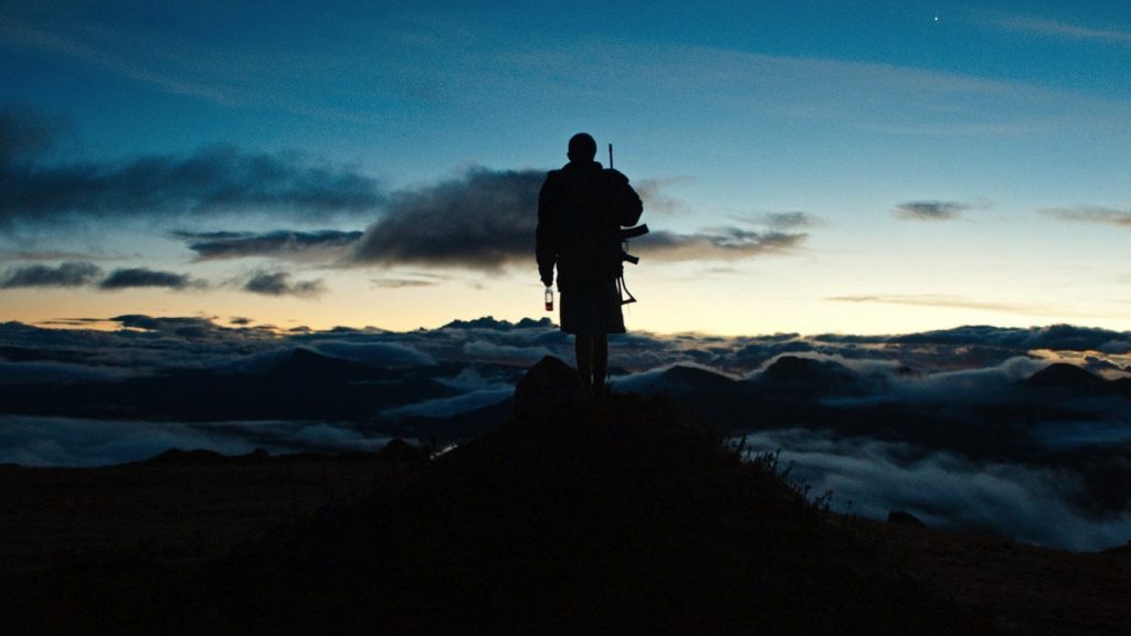 Silueta de persona de pie en cumbre montañosa al atardecer con nubes bajas y cielo azul gradiente