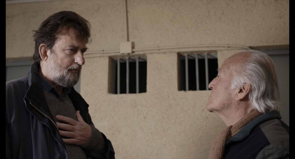 Dos hombres conversan en una celda de prisión; uno viste chaqueta negra y el otro es mayor con cabello gris