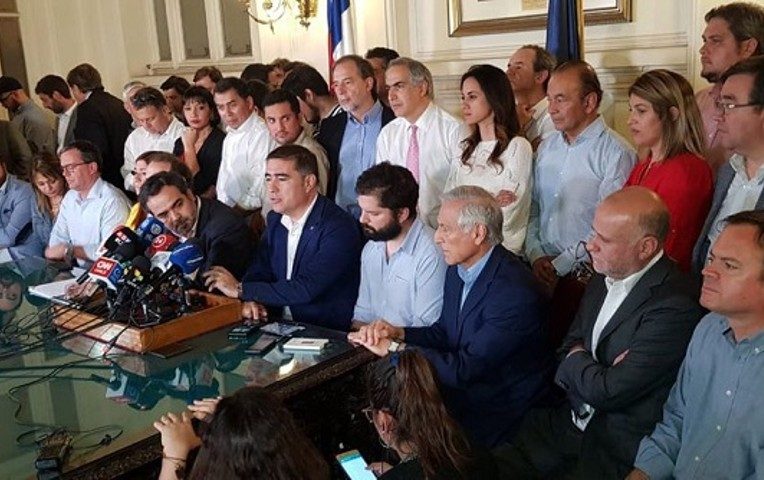 Grupo de personas en acto oficial en sala formal, con micrófono y prensa presente durante evento político