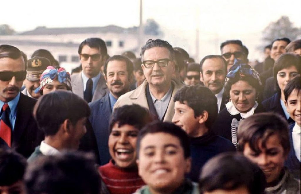 Salvador Allende en medio de una multitud, rodeado de seguidores y colaboradores en un acto público