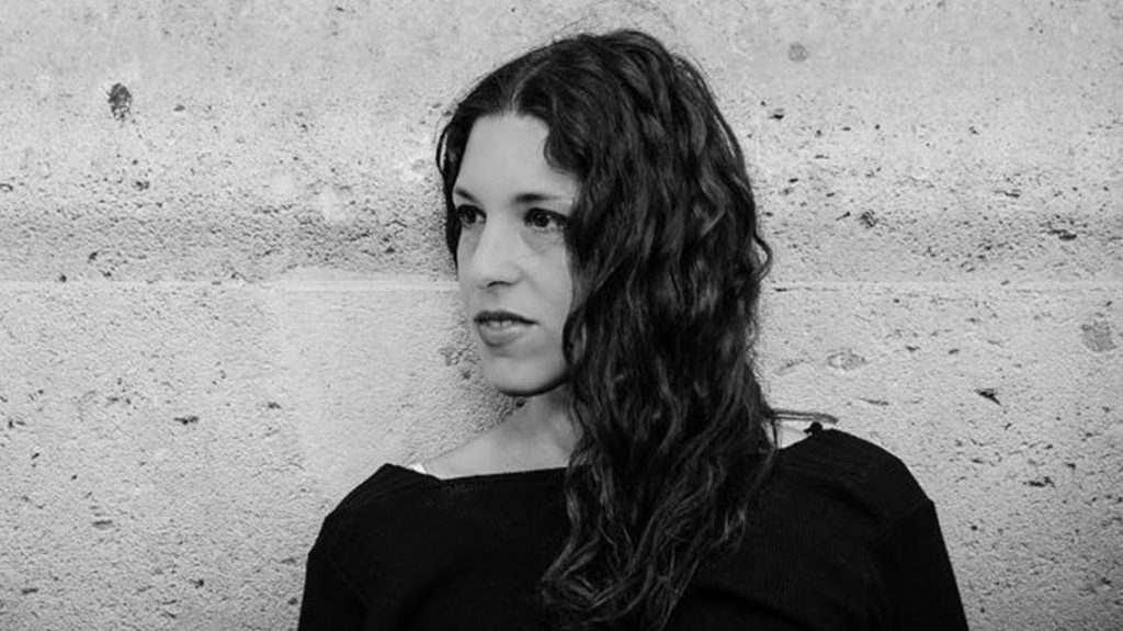 Mujer de cabello largo y oscuro ondulado, vistiendo ropa negra, posada contra pared de hormigón gris en fotografía