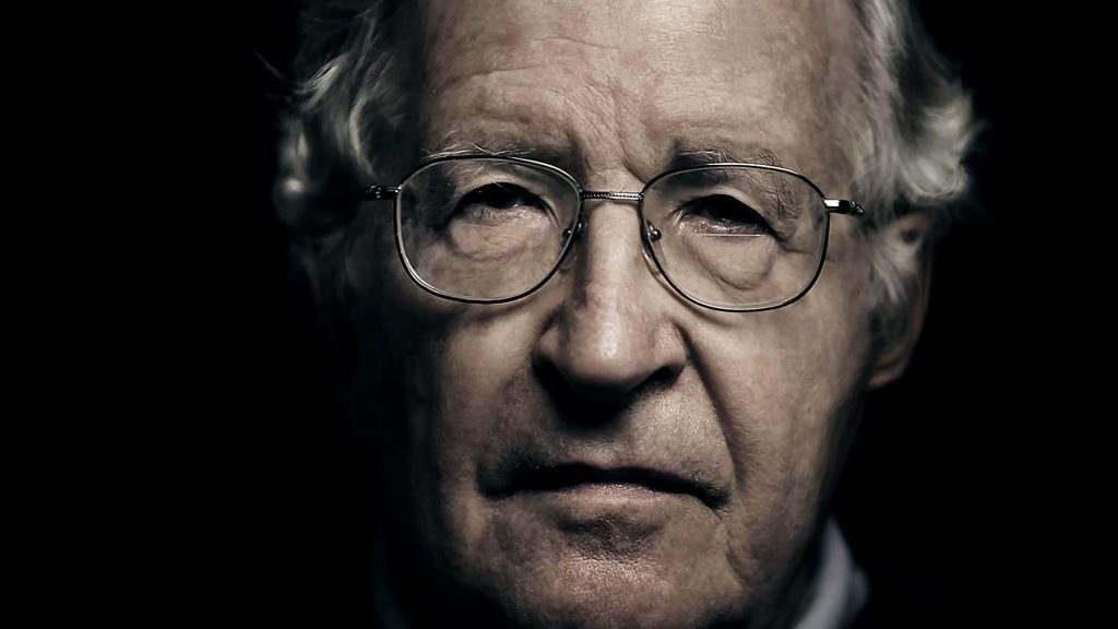 Noam Chomsky, hombre mayor con gafas redondas y cabello gris, retrato en fondo oscuro