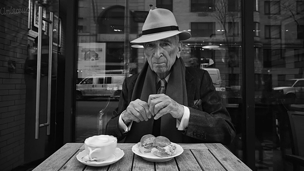 Gay Talese con sombrero y abrigo oscuro sentado a una mesa, con platos frente a él, en blanco y negro