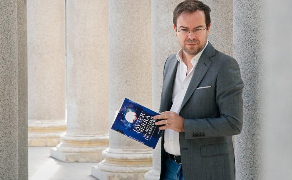 "El mensaje de pandora": Una reflexión de cara al futuro pospandémico Javier Sierra sostiene un libro azul junto a columnas clásicas, vistiendo traje gris oscuro