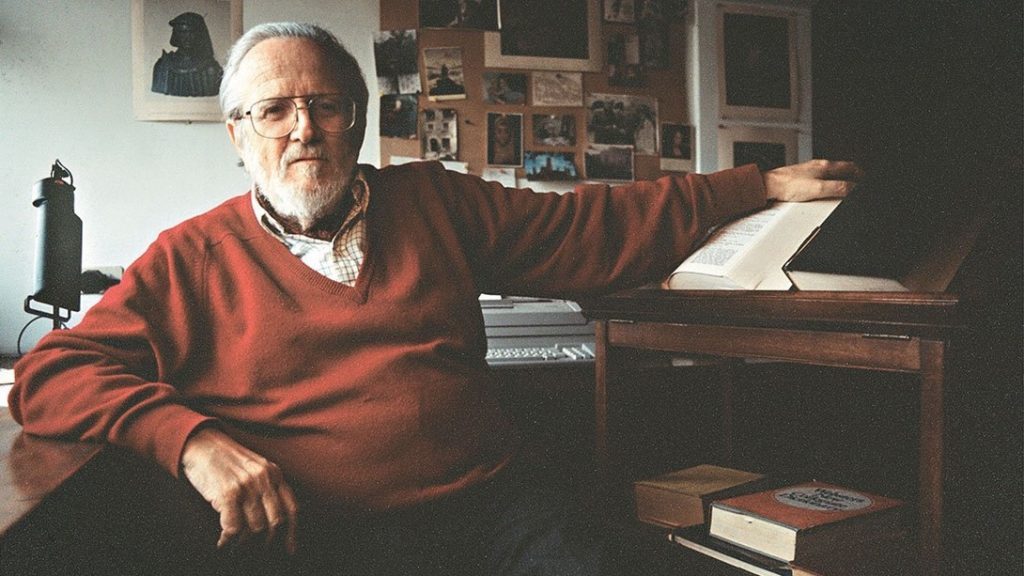 Escritor Gabriel García Márquez en su estudio, sentado ante escritorio con libros y papeles, vistiendo suéter rojo