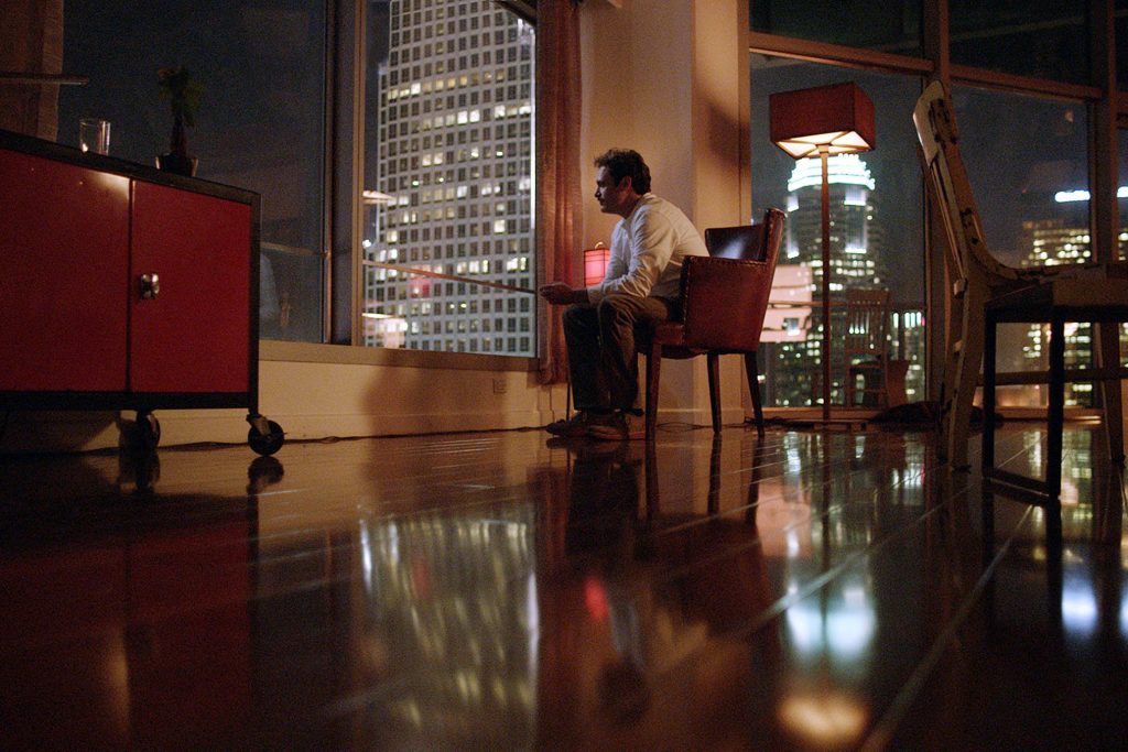 Hombre sentado en habitación minimalista de noche, frente a pantalla luminosa, rodeado de edificios iluminados al fondo
