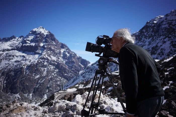 Patricio Guzmán filma con cámara de cine en la cordillera nevada, montañas rocosas al fondo bajo cielo azul
