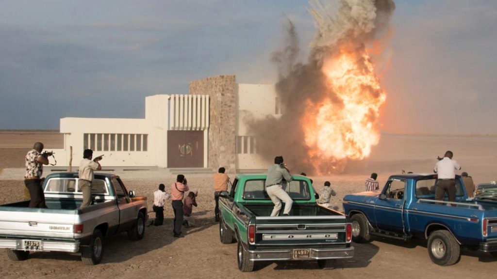 Escena de acción con vehículos antiguos y explosión de fuego en desierto árido, ambiente de violencia y caos