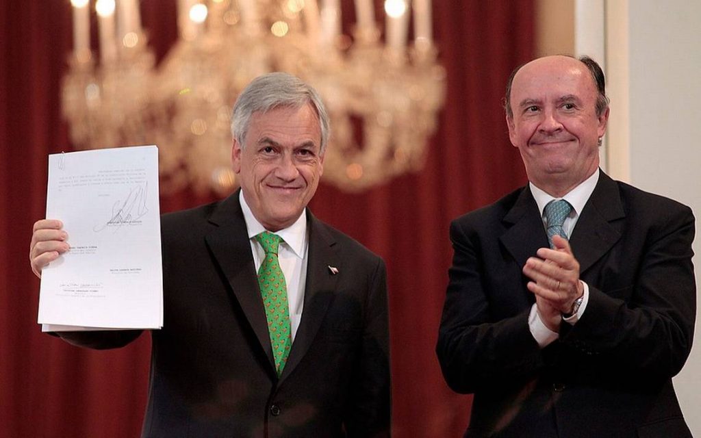 Sebastián Piñera sostiene documento mientras Cristián Larroulet aplaude, en acto oficial con decoración de candelabro