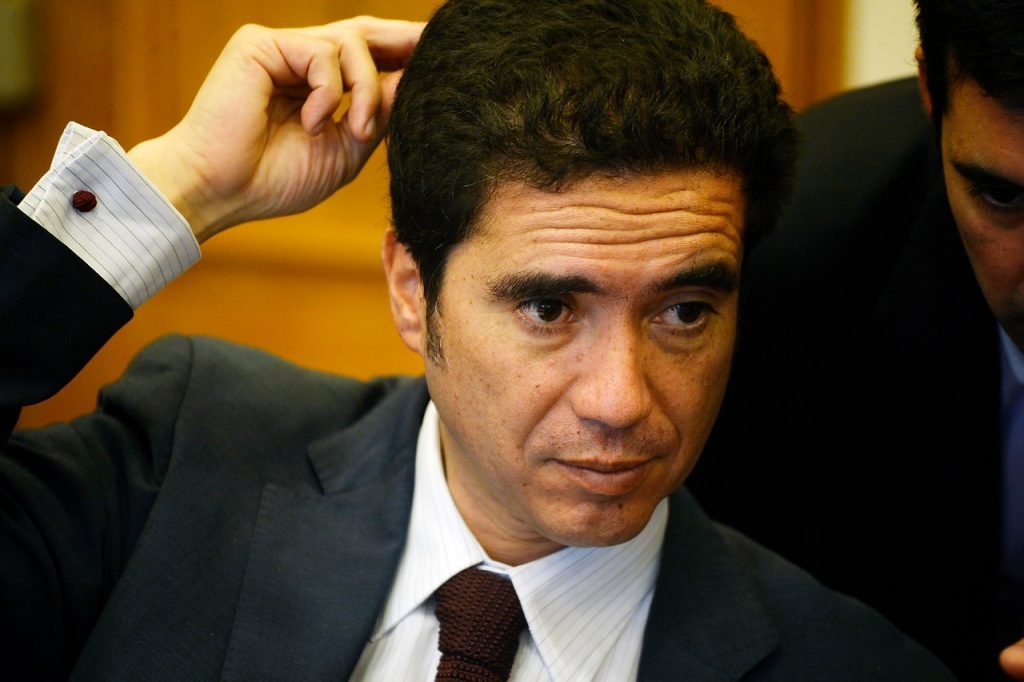 Hombre de traje oscuro y corbata roja en interior formal, con la mano en la cabeza, expresión seria