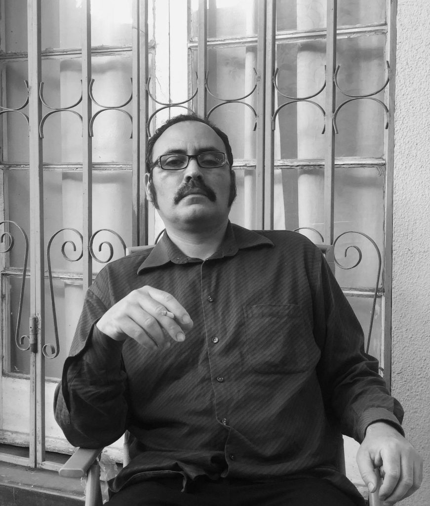 Poeta Rodrigo Verdugo en fotografía blanca y negra, con gafas y bigote, sentado frente a ventana de cristales