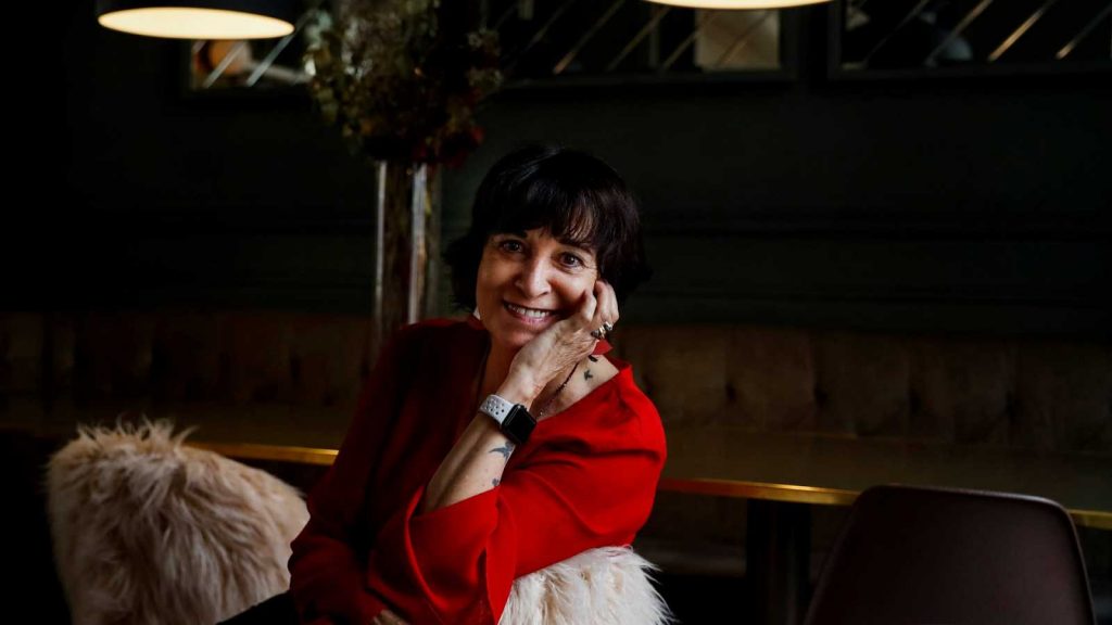 Rosa Montero, escritora española, sonríe mientras sostiene un cigarro, vistiendo chaqueta roja, en ambiente interior