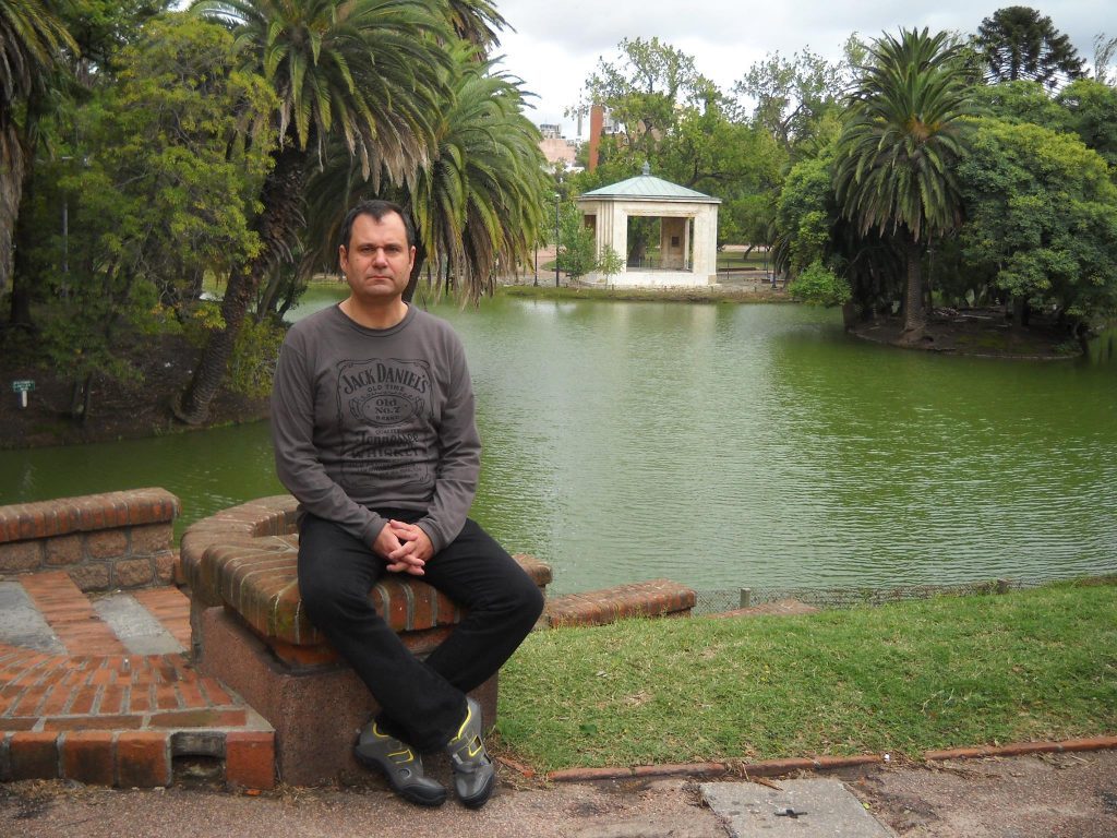 Aníbal Ricci sentado junto a un estanque con jardín y construcción clásica al fondo